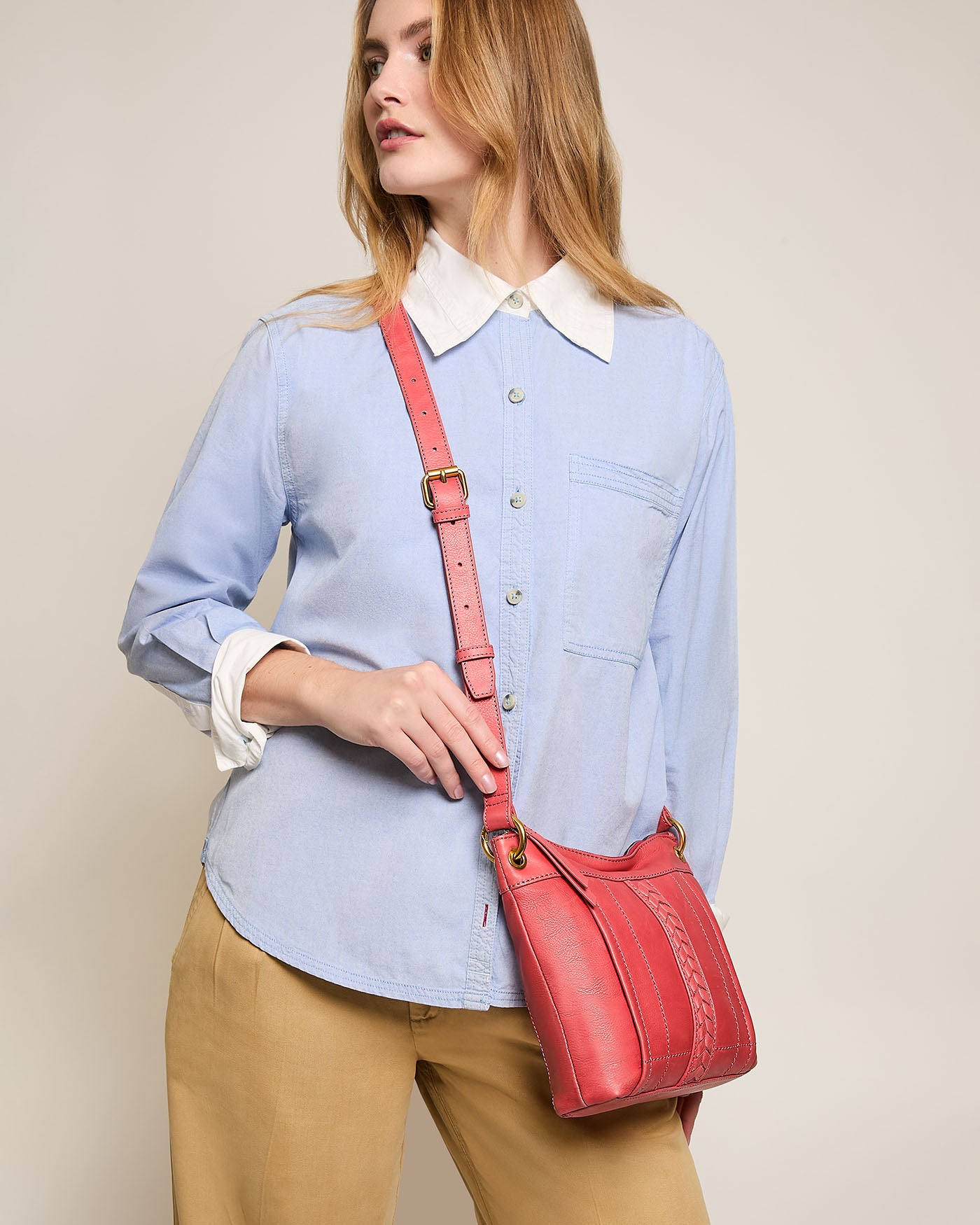 A person with long hair is wearing a light blue button-up shirt and beige pants, holding a red Sherryl Crossbody bag from American Leather Co. They tilt their head slightly while looking off to the side against a plain, beige background.