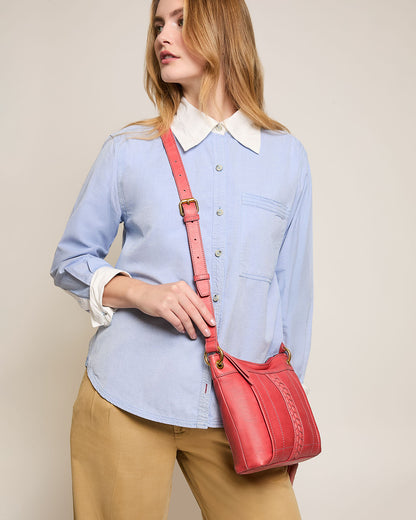 A person with long hair is wearing a light blue button-up shirt and beige pants, holding a red Sherryl Crossbody bag from American Leather Co. They tilt their head slightly while looking off to the side against a plain, beige background.