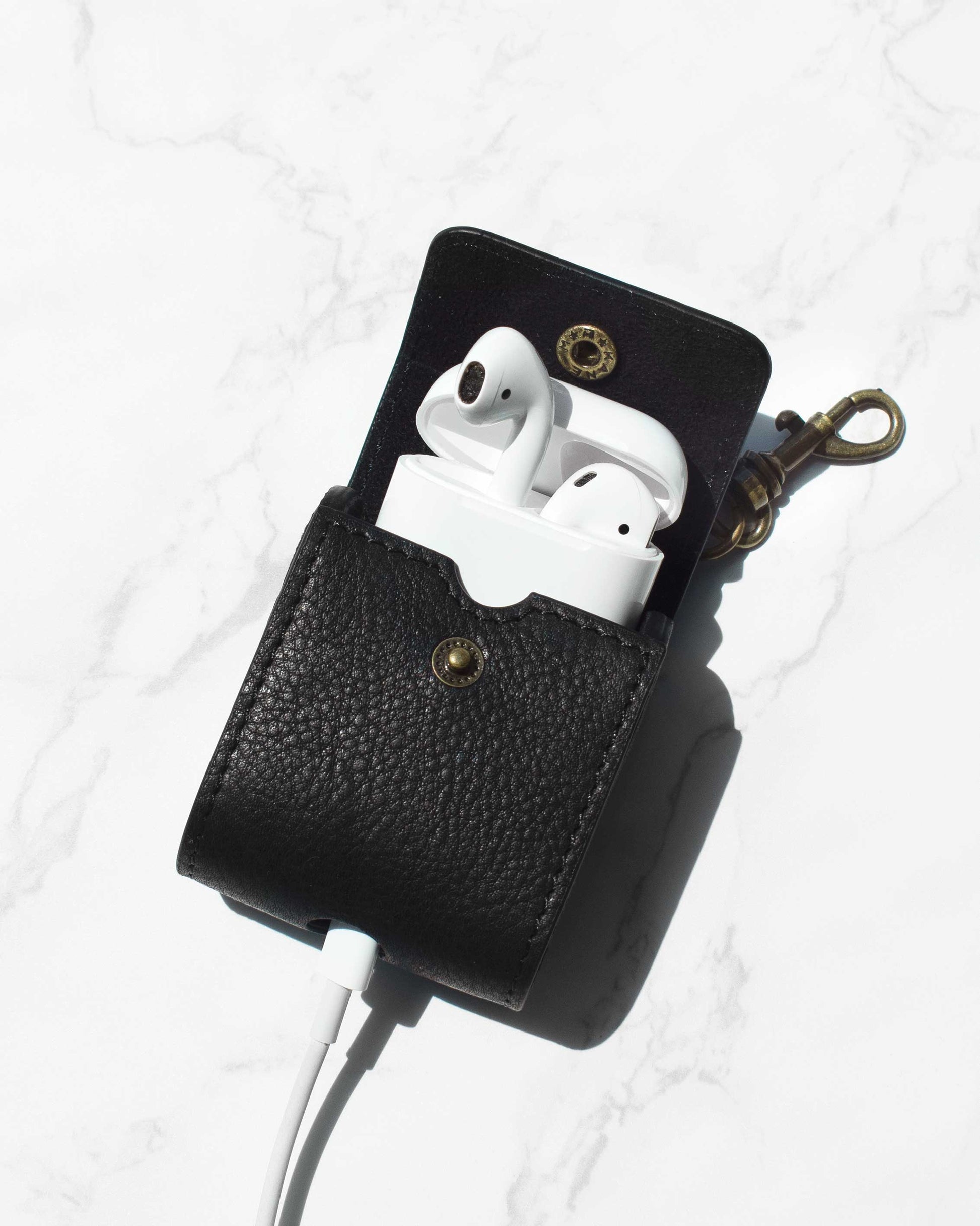 Flagstaff Airpod Case by American Leather Co.: White wireless earbuds encased in a genuine black American leather case featuring a snap button, displayed on a marble surface. The design includes an integrated charger cord opening and an attached AirPods bag charm metal clip.