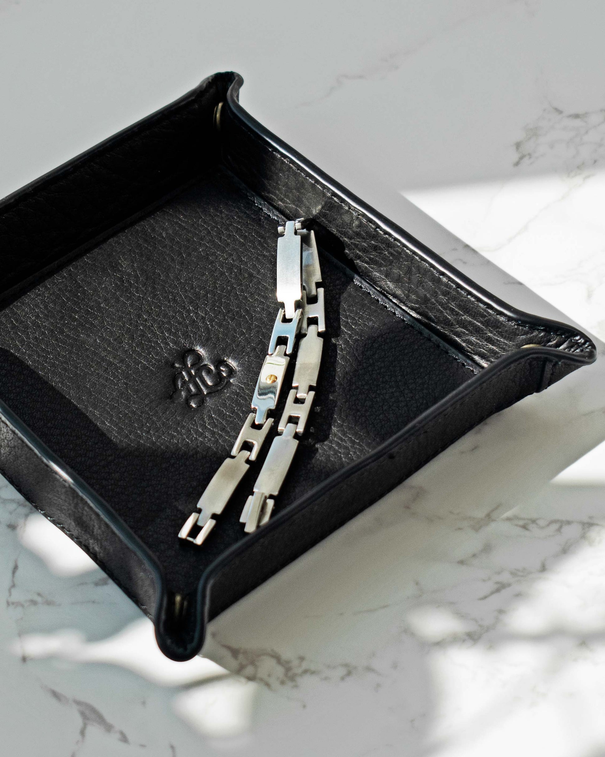 A silver link bracelet rests in an open Manchester Leather Tray by American Leather Co. on a white marble surface. Sunlight casts soft shadows across the scene, highlighting the genuine leather and the elegance of antiqued hardware.