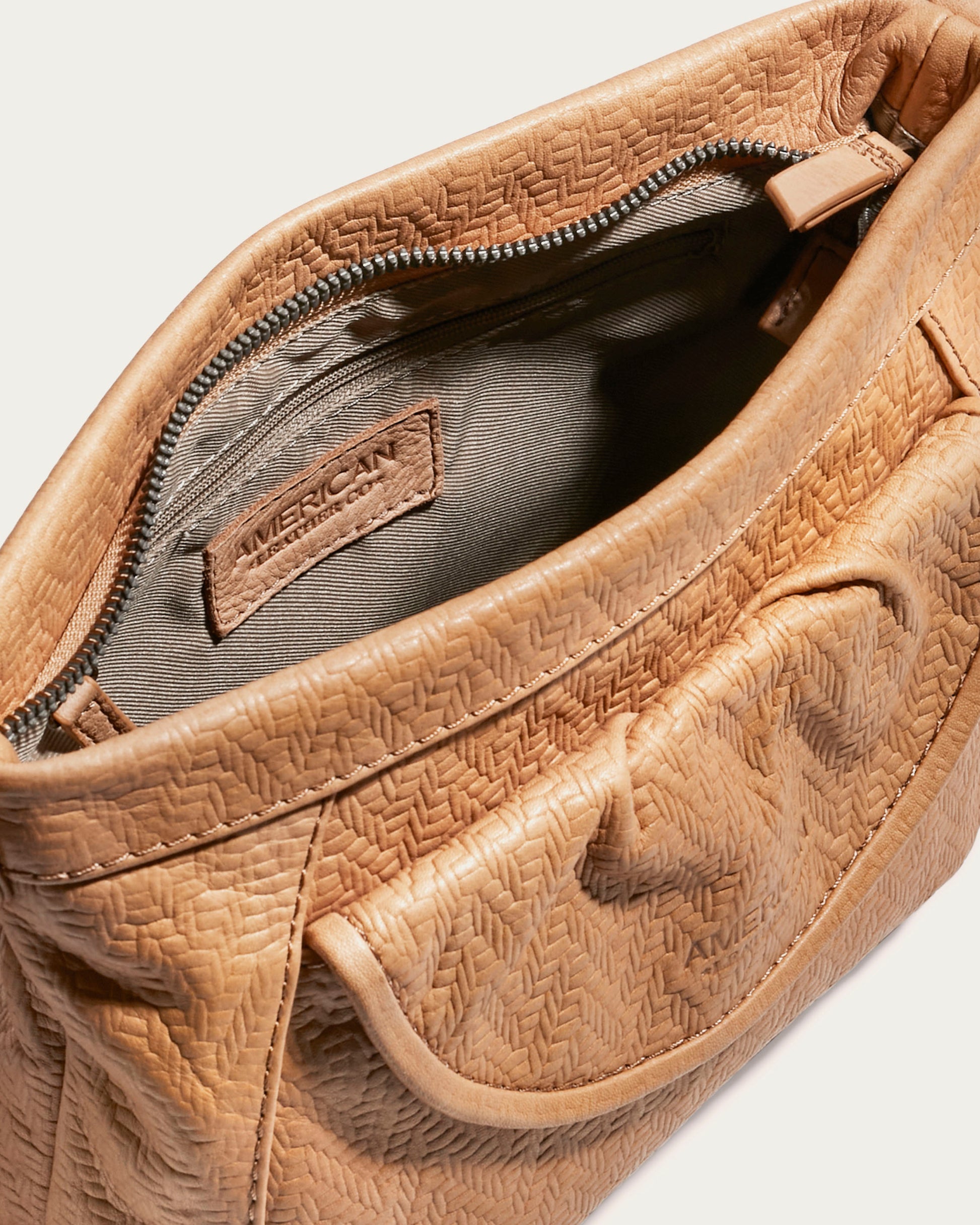 Close-up of an open Reed Crossbody handbag in beige, showcasing a textured woven pattern with glove leather accents. The interior is lined with gray fabric and includes a visible zipper along with a tag labeled "American Leather Co." The exterior features a front pocket, detailed stitching, and an optional crossbody strap for added versatility.