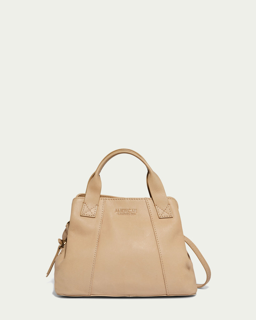 Introducing the Ada Triple Entry Crossbody by American Leather Co. - a beige genuine leather satchel featuring short handles and a detachable shoulder strap. This bag showcases a simple, elegant design with the branded logo embossed on the front, set against a plain off-white background.