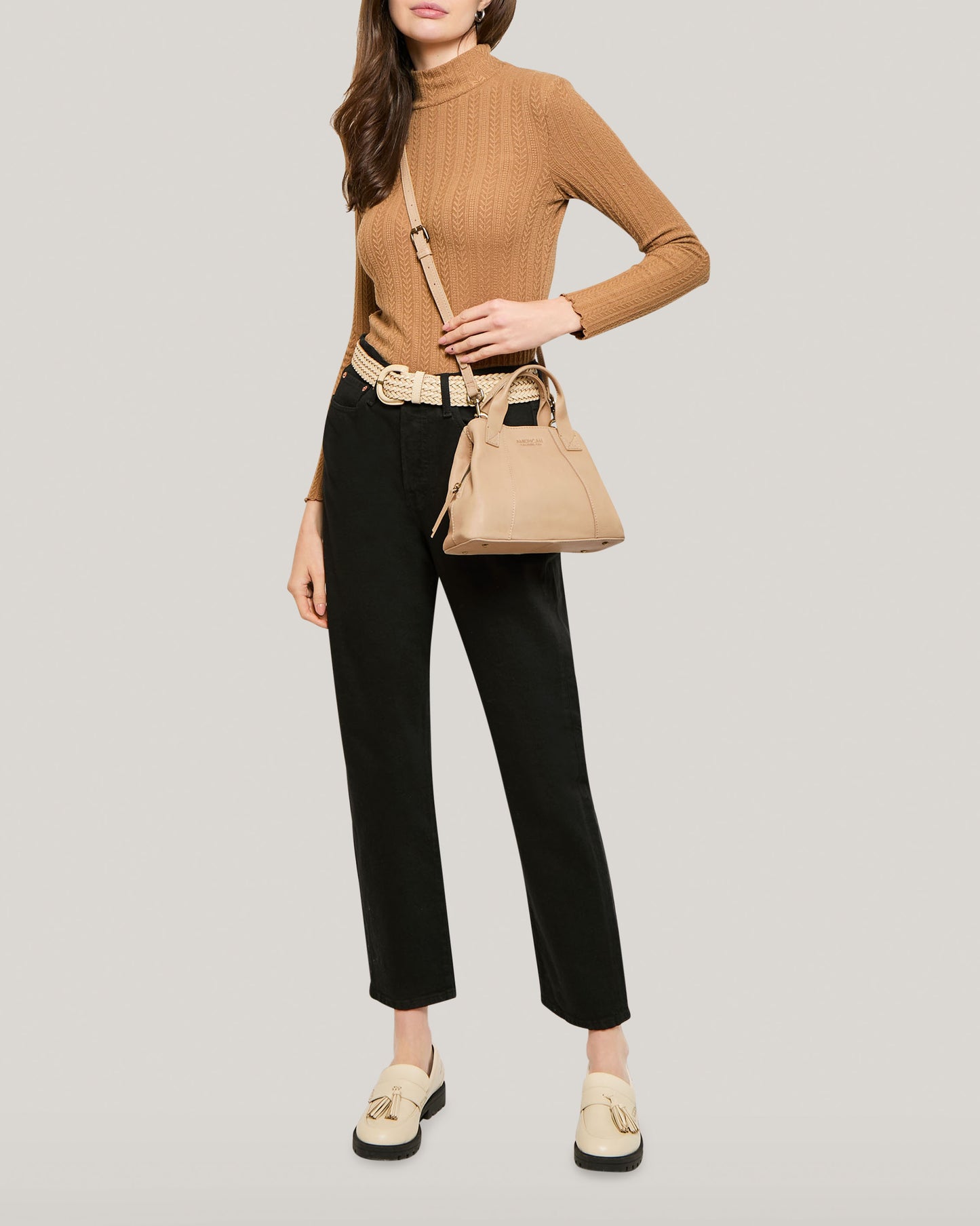 A person is wearing a long-sleeved camel-colored turtleneck sweater, black high-waisted trousers, and cream-colored loafers. They are carrying an Ada Triple Entry Crossbody by American Leather Co. made of genuine leather, and have a beige belt around their waist. The background is a plain light gray.