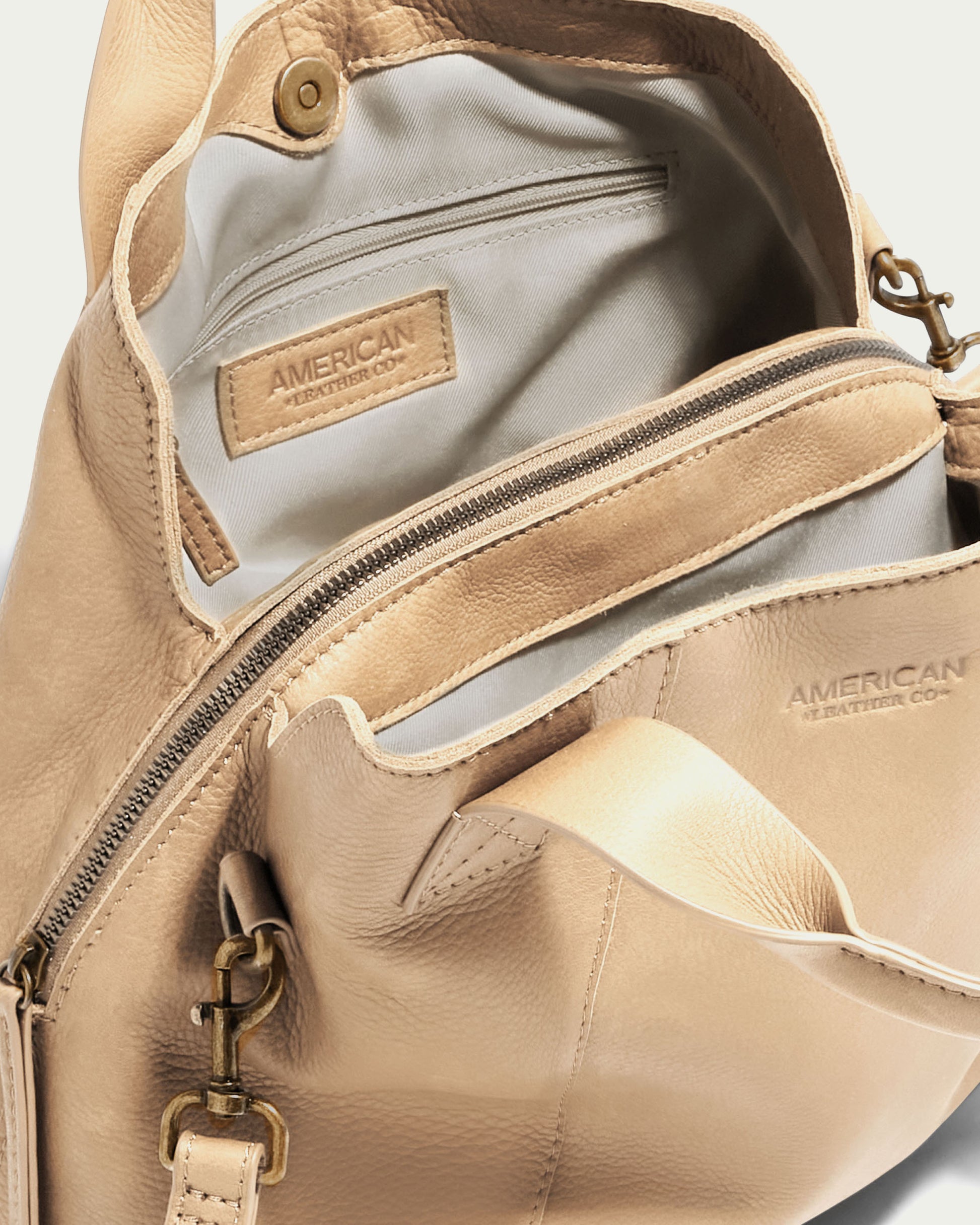 The Ada Triple Entry Crossbody, a beige leather bag from American Leather Co., is displayed. The interior is light gray and includes a zippered pocket along with a fabric tag reading "AMERICAN LEATHER CO." A similar tag is positioned on the exterior above a large zippered pocket.