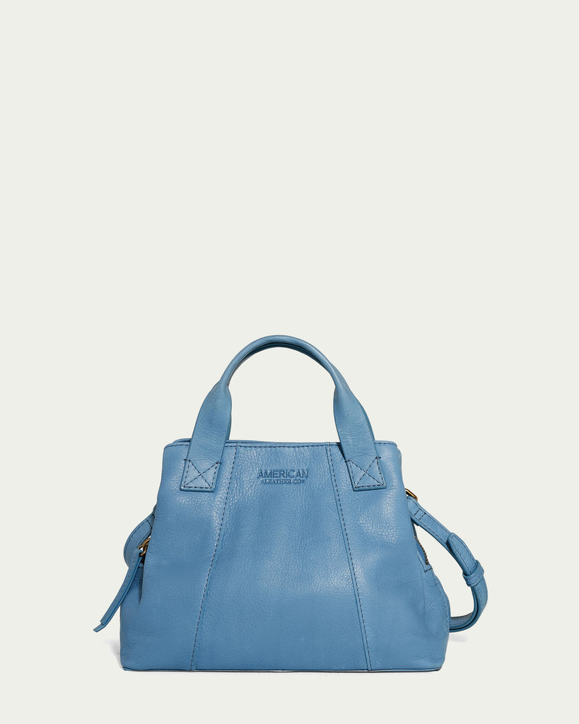 The Ada Triple Entry Crossbody, a light blue genuine leather handbag from American Leather Co., is showcased against a plain background. It boasts two sturdy top handles and includes a detachable crossbody strap. The front is elegantly embossed with "American Leather Co." and its triple entry design gives it a chic, structured square shape.