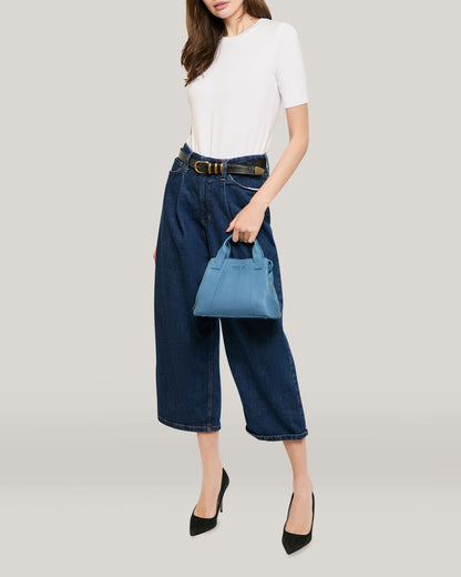 A person with long hair is wearing a white short-sleeve top, dark blue wide-leg jeans, and black heels. They are holding a small Ada Triple Entry Crossbody by American Leather Co. and standing against a plain light-colored background. Their face is not visible.