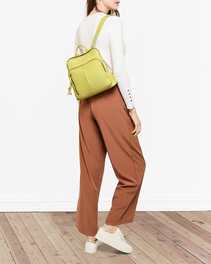 A person with long hair is posing against a plain background. They are wearing a white long-sleeve top, brown pants, and white sneakers. They have the Cleveland Midsize Backpack by American Leather Co., featuring a front zip compartment, on their back and are standing on a wooden floor.