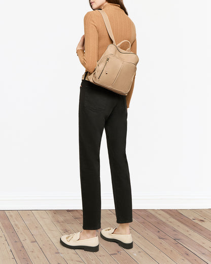 A person in a tan sweater and black pants is on a wooden floor. They carry the stylish American Leather Co. Cleveland Midsize Backpack with a convenient zip compartment and wear matching beige loafers. The background is plain white.