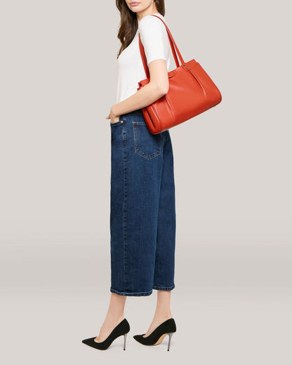 A person is standing against a plain background, wearing a white top, blue high-waisted jeans, and black high heels. They have the Layton Satchel by American Leather Co., a red leather bag with multiple interior pockets, slung over their shoulder. Only part of the person's face is visible.