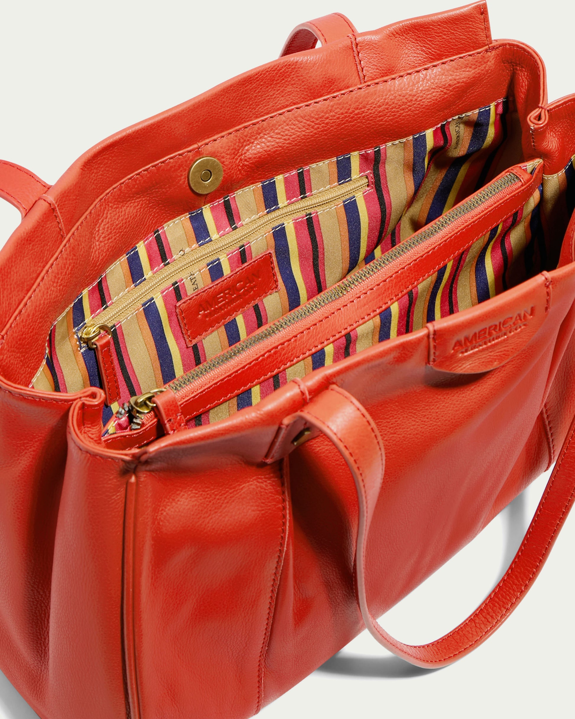 The Layton Satchel by American Leather Co. is a bright red leather tote bag featuring gold zippers and button closures. The interior, lined with vibrant, multicolored striped fabric, offers multiple interior pockets and a center zipper compartment for organization. This bag boasts sturdy handles and a clean finish.