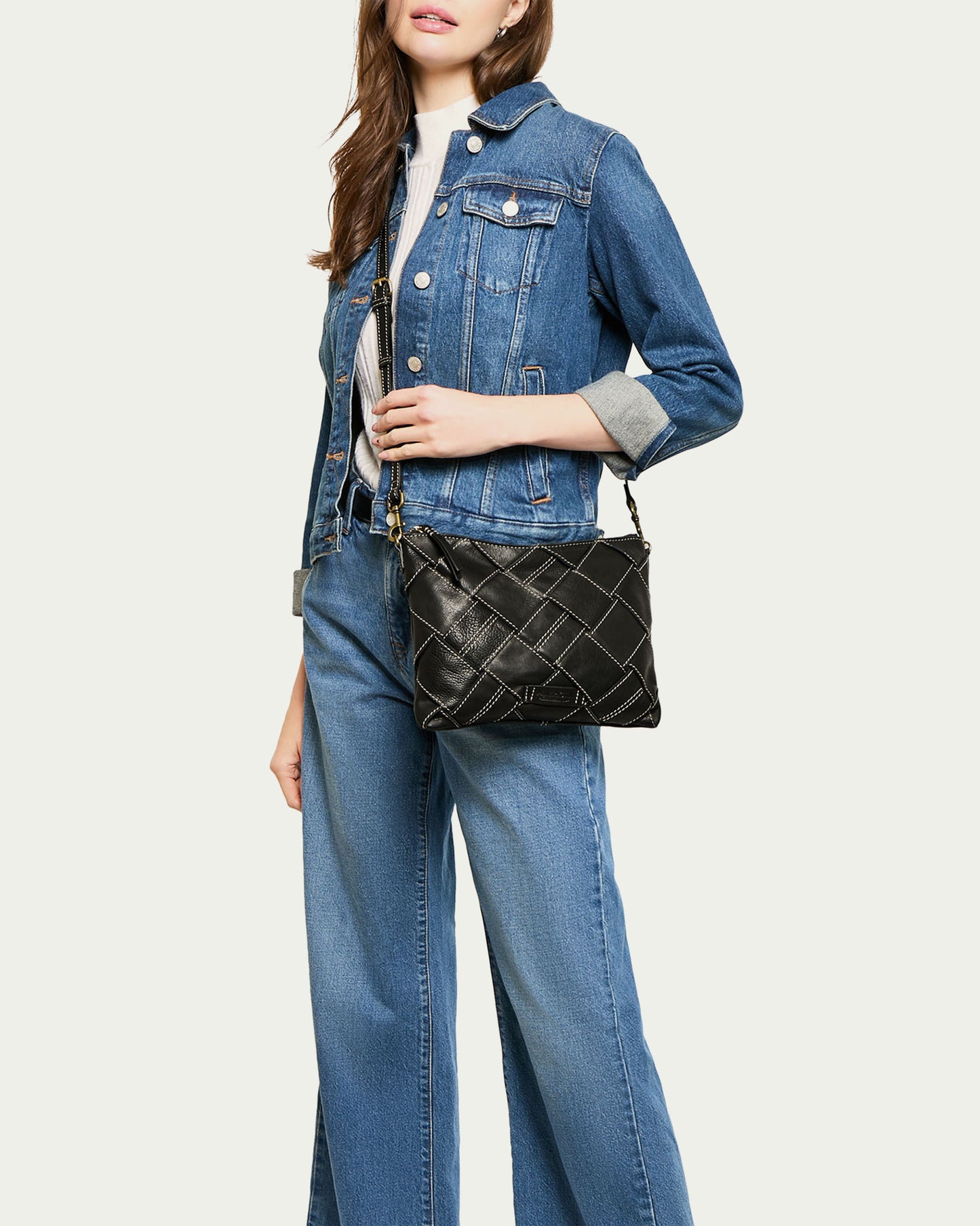 A woman dressed in a denim jacket and blue jeans holds an American Leather Co. Mercer Woven Convertible Clutch with a chain strap. This outfit embodies a casual yet fashionable style, perfectly accented by the chic accessory against a plain light background.