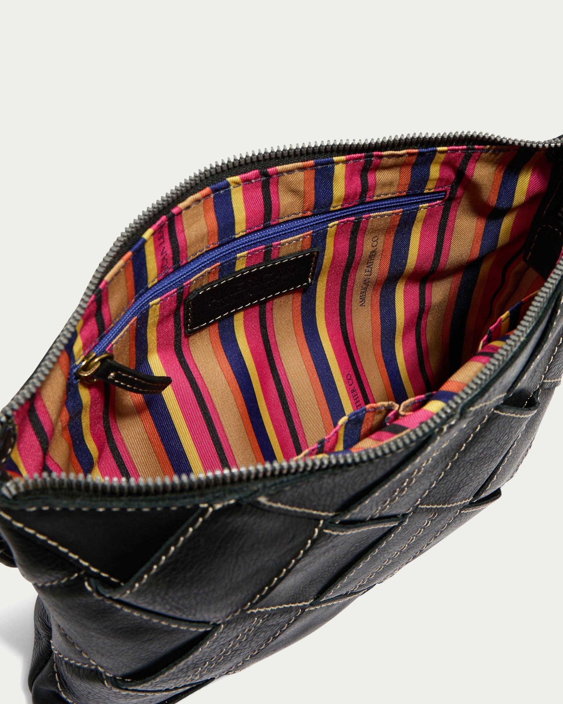 The Mercer Woven Convertible Clutch by American Leather Co. is a black woven leather handbag with visible stitching that reveals a vibrant, striped lining in pink, yellow, blue, and orange. This versatile piece includes an interior zippered pocket and a spacious main compartment.