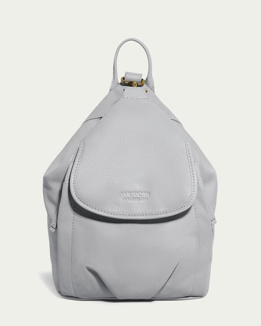 Introducing the Pierce Backpack by American Leather Co., a light gray genuine leather design featuring a front flap with subtle branding centered on it. This backpack includes a top handle and adjustable shoulder straps, set against a plain light-colored background.