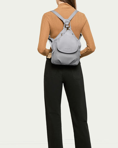 A person in a beige turtleneck sweater and black pants is seen from behind, carrying the Pierce Backpack by American Leather Co., which features genuine leather adjustable straps in a light gray leather design. The background is plain and light-colored.