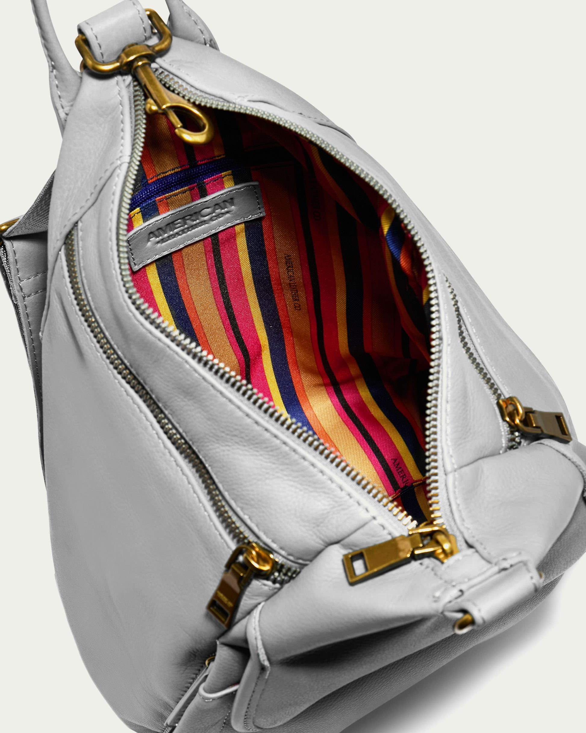 The Pierce Backpack by American Leather Co. features a gray leather exterior and opens to reveal a colorful striped interior in shades of red, orange, yellow, and purple. It is accented with gold zippers and hardware and comes with adjustable straps for versatile wear.