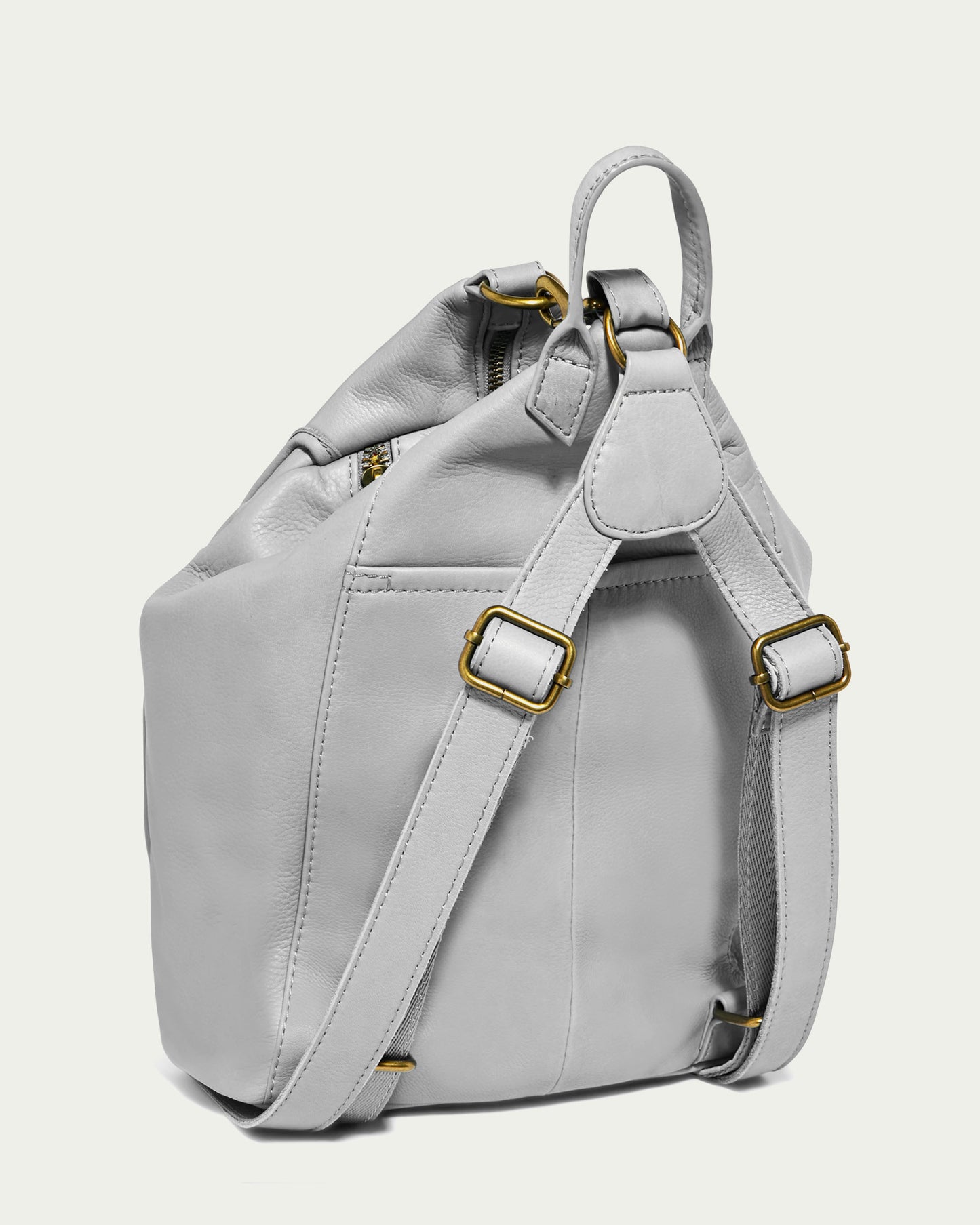 The Pierce Backpack by American Leather Co. is displayed from the side, showcasing its stylish design. Made from genuine leather in a light grey shade and accented with gold-colored hardware, it features adjustable straps and a convenient top handle. Its elegant style makes it perfect for both casual and formal occasions.