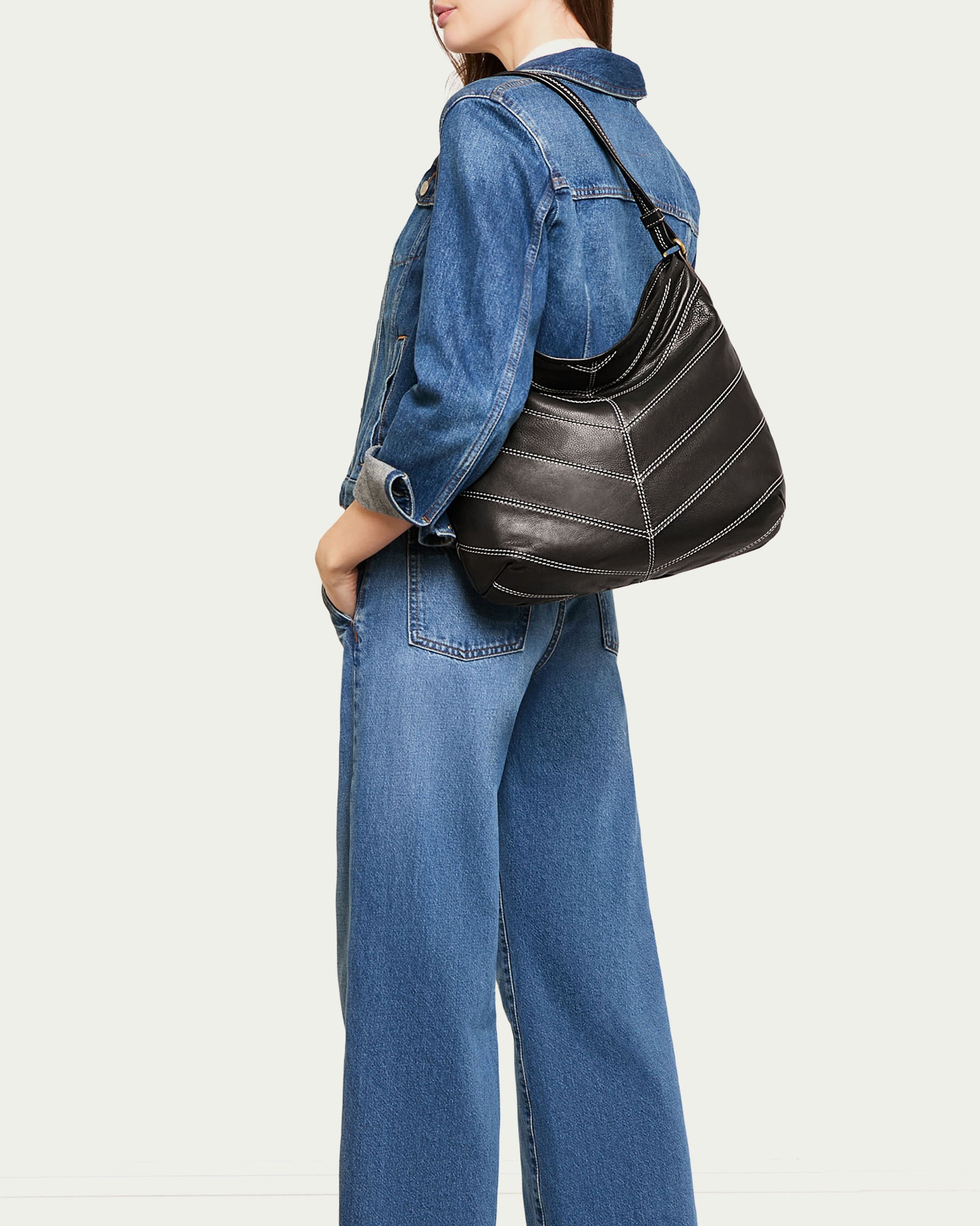 A person wearing a blue denim jacket and jeans holds the Raleigh Ziptop Patchwork Hobo by American Leather Co., a black, genuine leather bag. The background is plain off-white, and the person's head is not fully visible.