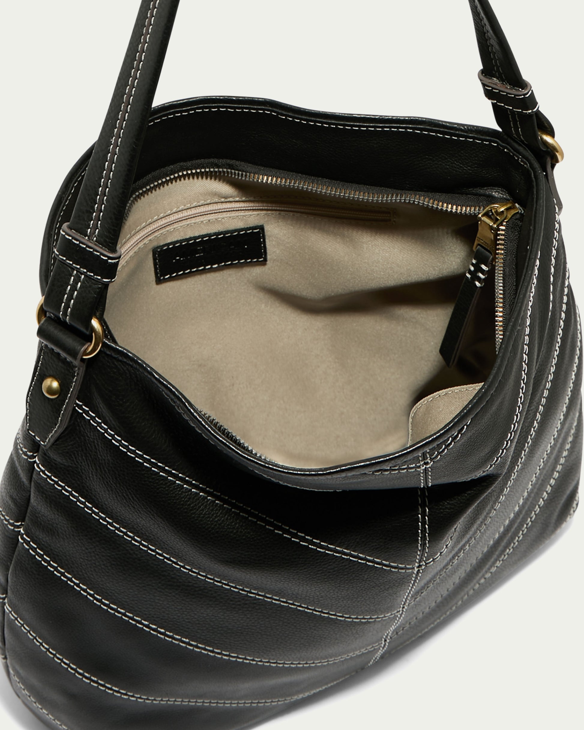 The Raleigh Ziptop Patchwork Hobo by American Leather Co. is an elegant open hobo bag crafted from genuine black leather, featuring striking white stitching that reveals a glimpse of its beige interior. This stylish accessory includes a single shoulder strap and a zippered inner pocket, all highlighted by its eye-catching chevron patchwork design.