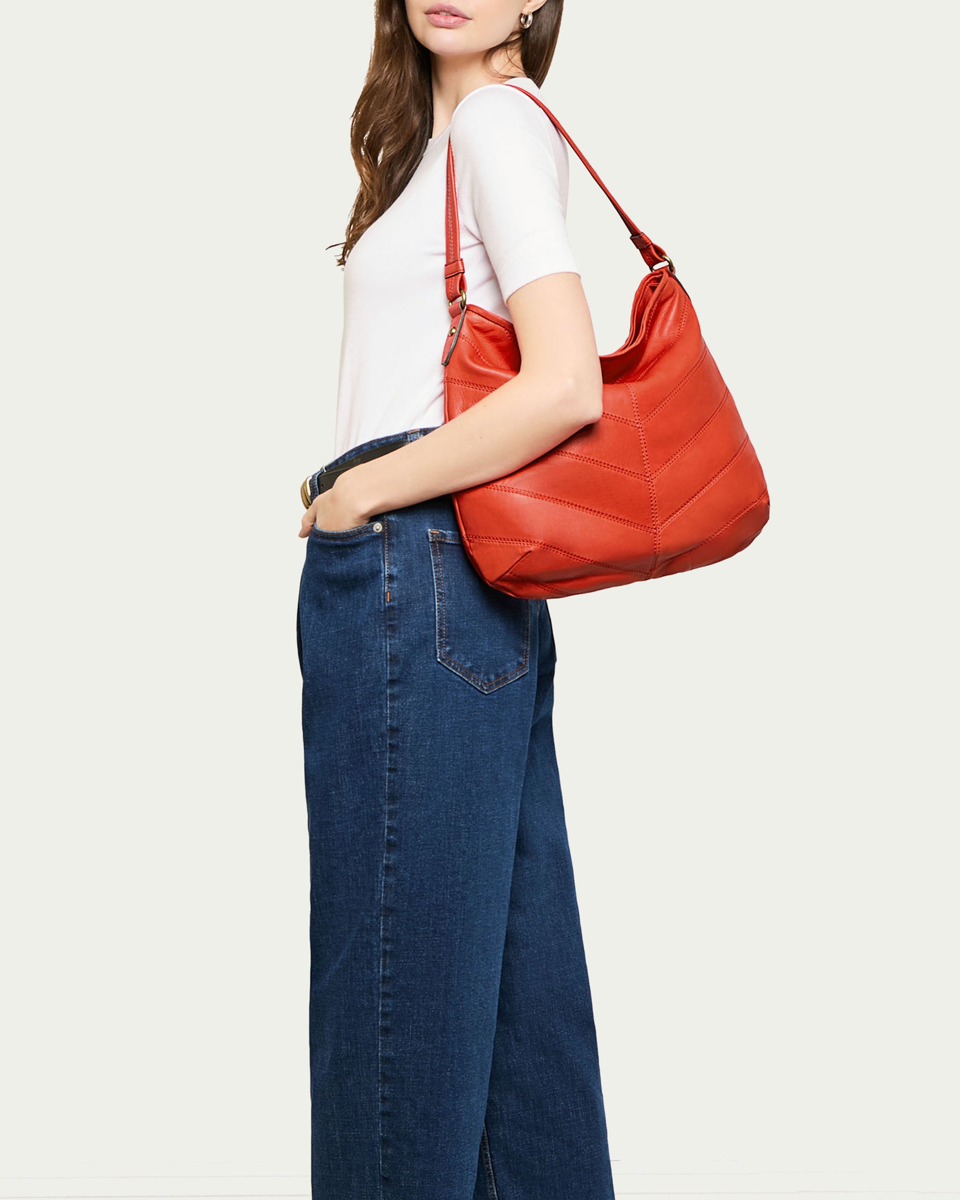 A person with long hair models the American Leather Co. Raleigh Ziptop Patchwork Hobo, a red genuine leather shoulder bag. They wear a white short-sleeved top and high-waisted blue jeans. The plain background highlights the bag's chevron patchwork design and zip-top closure, adding an elegant touch to the ensemble.