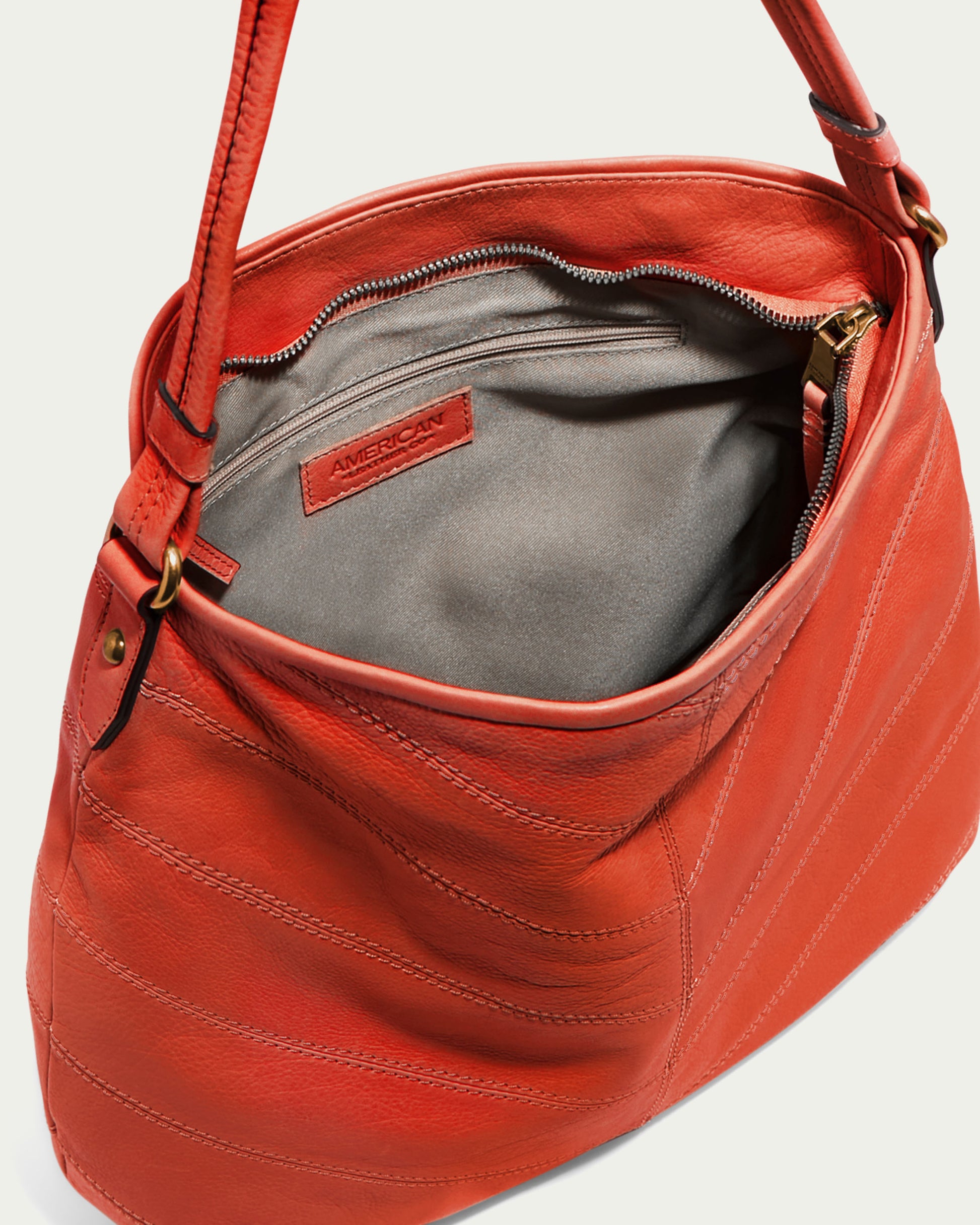 The Raleigh Ziptop Patchwork Hobo by American Leather Co. showcases a stylish chevron patchwork design from a top-down perspective, revealing its large orange leather body with two straps. Inside, the bag features a gray interior with a zippered pocket and an "American Leather Co." tag. The genuine leather exterior is adorned with stitching details and offers a secure zip top closure.