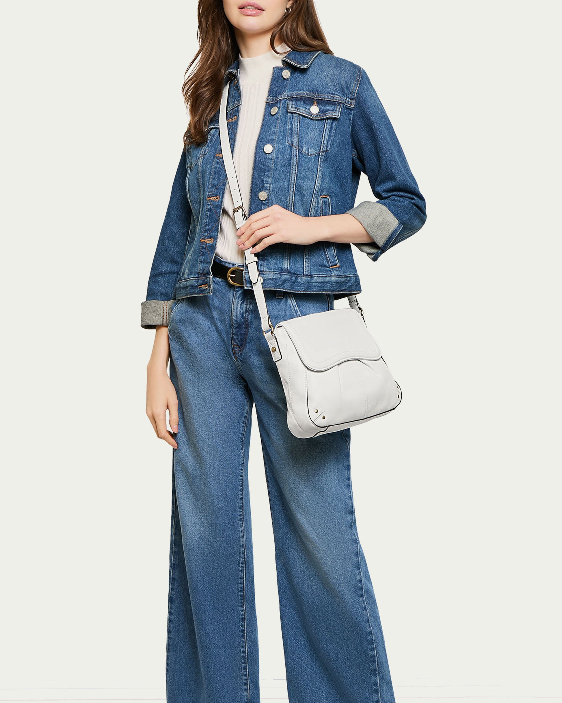 A woman dressed in a denim jacket over a white top and wide-leg jeans is carrying the Ria NS Crossbody bag by American Leather Co., which features an adjustable strap. The backdrop is a simple light shade.