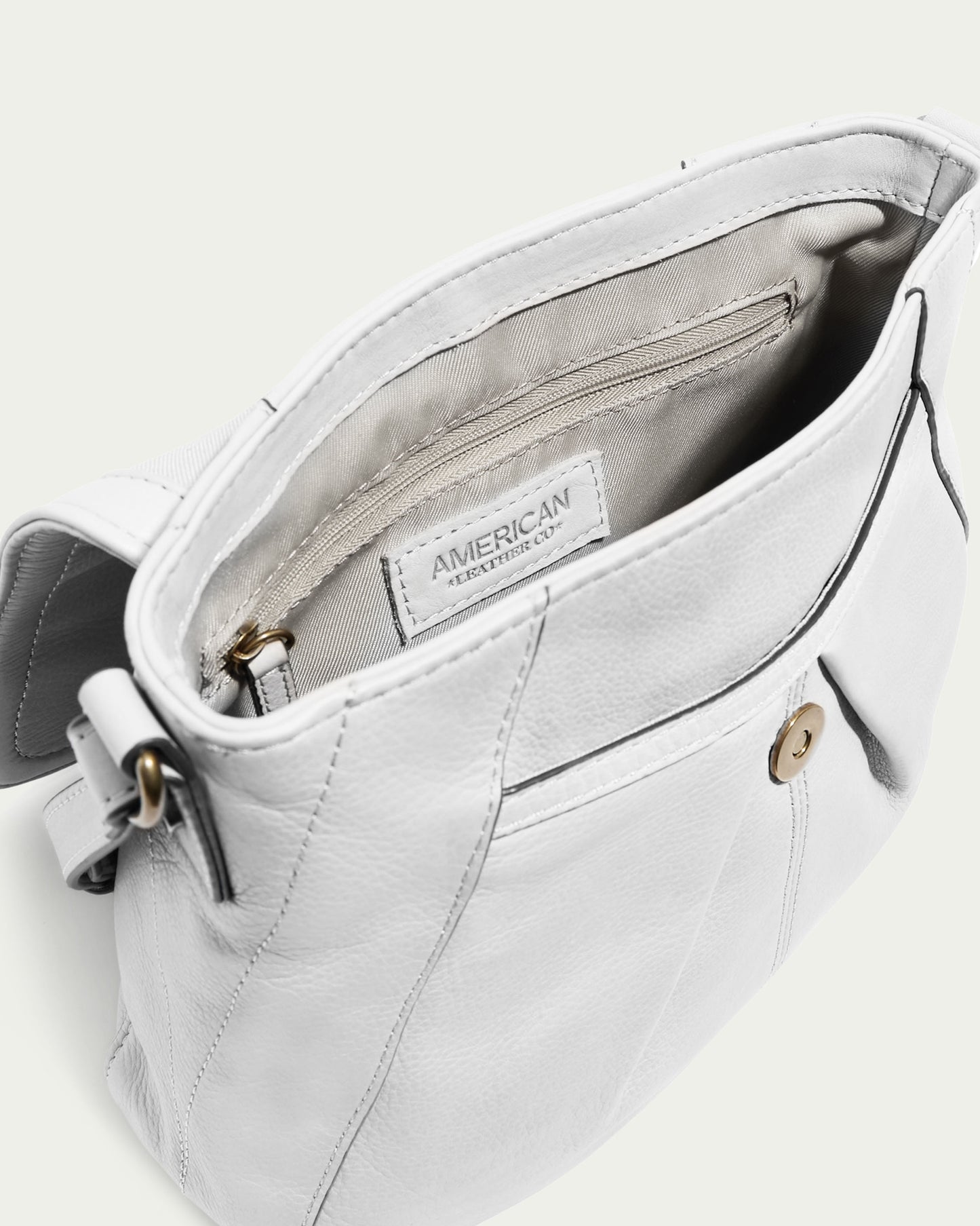 The Ria NS Crossbody by American Leather Co. is a white genuine leather handbag with an open top that reveals a fabric interior and a zippered pocket. It features a brass button closure on the front and comes with an adjustable strap, making it perfect for use as a crossbody bag.