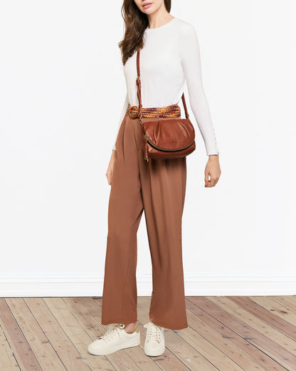 A person dressed in a white long-sleeve top, brown wide-leg pants, and white sneakers stands against a white background on a wooden floor. They are holding the Talya Double Entry Crossbody by American Leather Co., which features an adjustable strap.