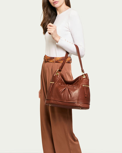 A person wearing a white long-sleeve shirt and brown pants is holding the Tenafly Hobo by American Leather Co., a stylish genuine leather handbag with gold accents and zipper pockets that complement the ensemble. The person stands against a light background.