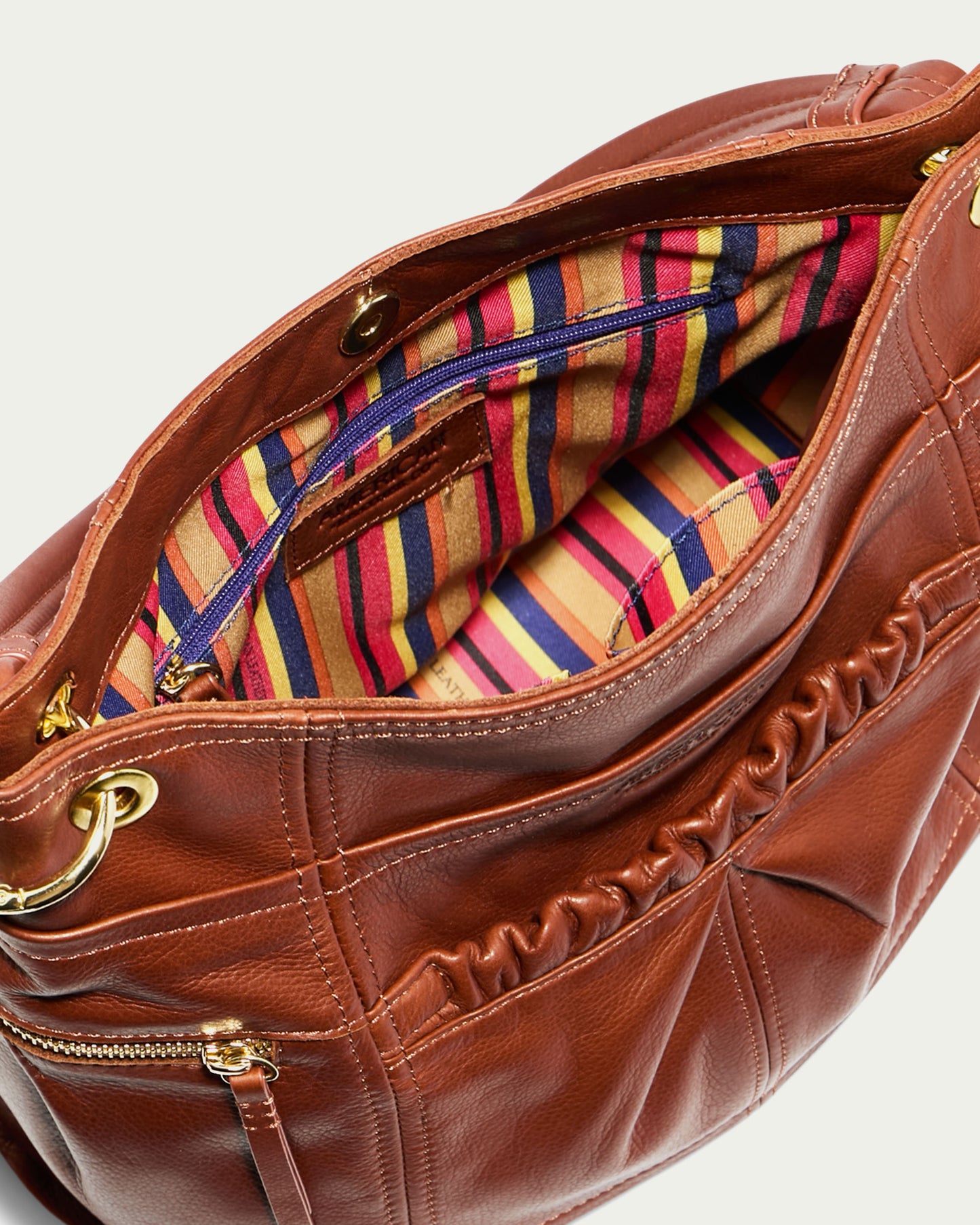 The Tenafly Hobo by American Leather Co. is a brown genuine leather bag with an open top, revealing a vibrant striped fabric lining inside. This stylish accessory is accented with gold hardware and includes a front zipper pocket, ideal for organizing essentials.