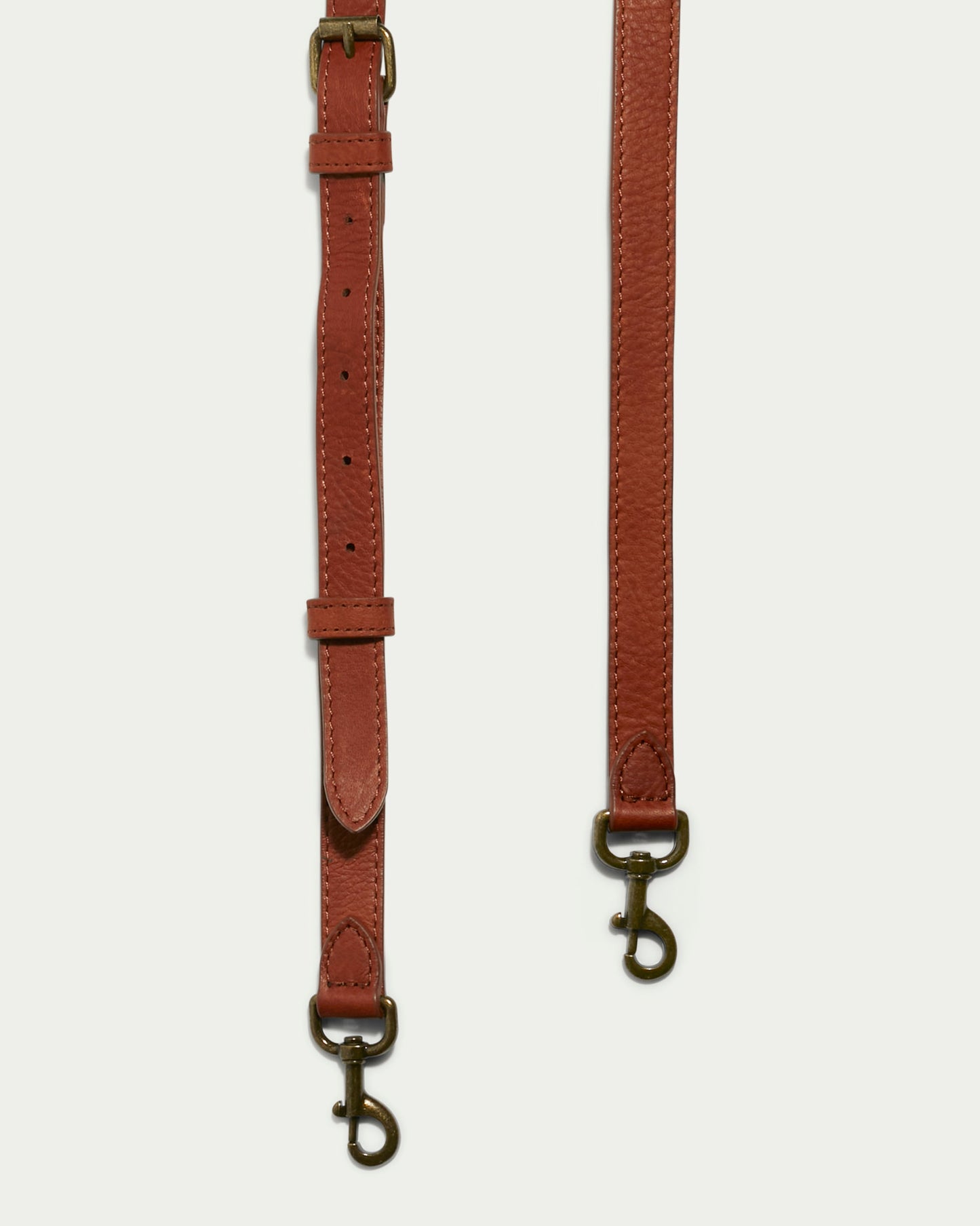 Two Tall Crossbody Straps in brown leather from American Leather Co. are displayed vertically against a light background. Both straps feature brass hardware and swivel snap hooks at the ends, with one strap offering multiple holes for adjustment.