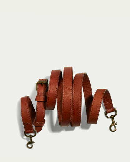 A product from American Leather Co. is displayed in a neat spiral on a light background. Identified as the Tall Crossbody Strap, this coiled brown leather dog leash features metal clasps on both ends and an adjustable buckle that emphasizes its elegant style.