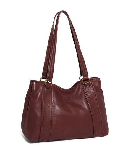 The Val Perfect Satchel by American Leather Co. is a maroon leather handbag with dual handles, antiqued gold hardware, and a soft, slightly textured finish. It features two vertical seams down the front, a slightly rounded top edge, and a spacious design suitable for everyday use. For added convenience, it includes a magnetic snap pocket.