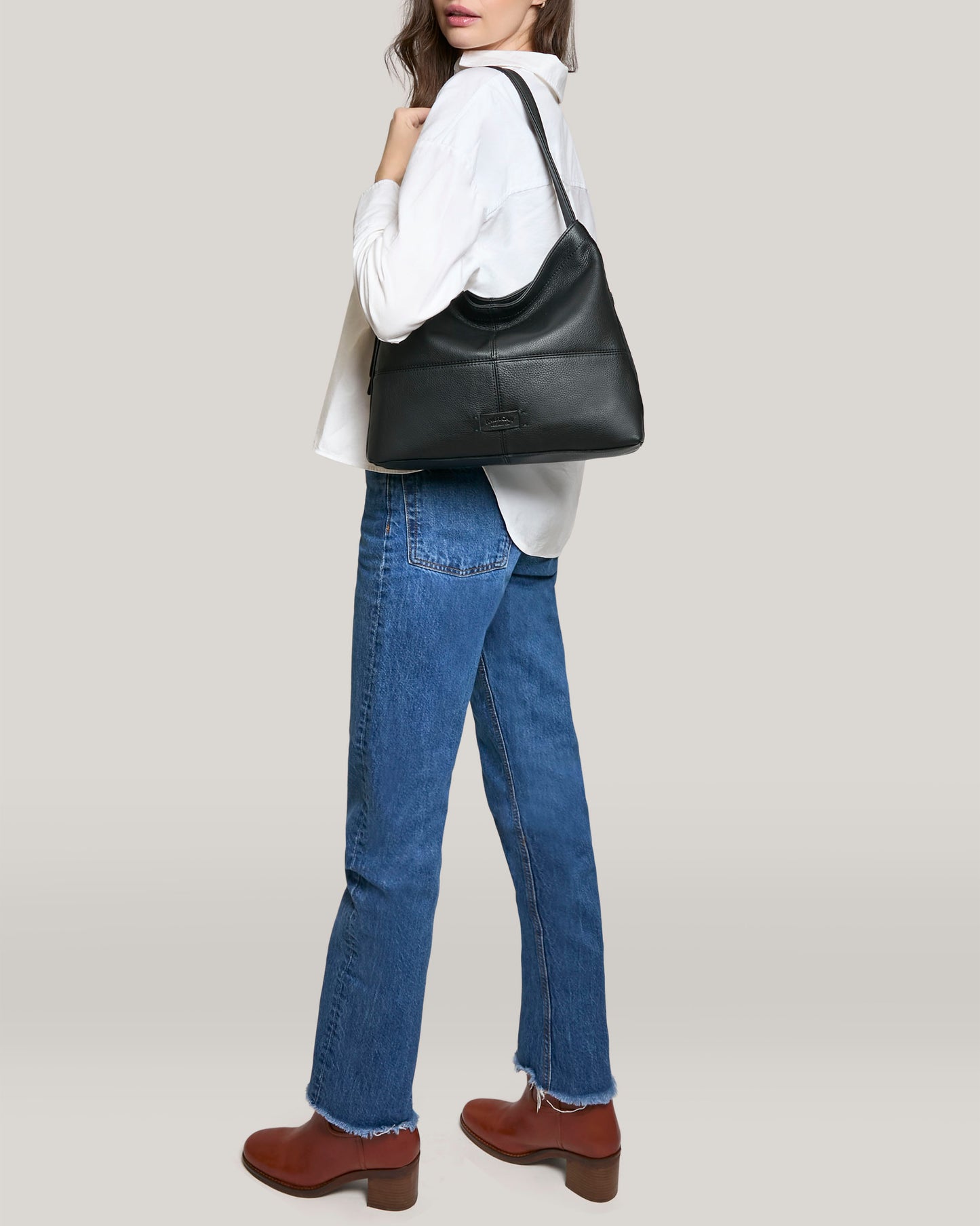 The individual poses sideways in a white shirt, blue jeans, and brown boots, holding a Virginia Hobo by American Leather Co. The artisanal contrast stitching on the black shoulder bag enhances elegance against the simple light gray background.