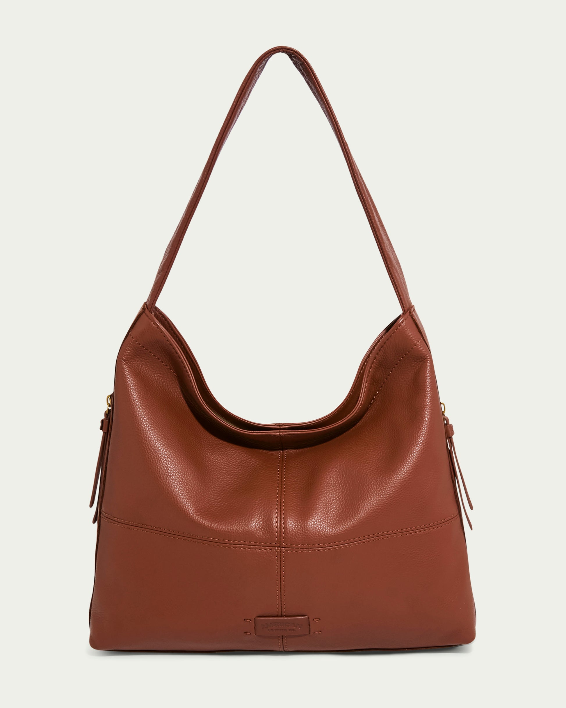 The Virginia Hobo by American Leather Co. is a brown leather handbag crafted from genuine American leather. It has a single shoulder strap, simple and elegant design, slightly curved top, and artisanal contrast stitching on a light background.