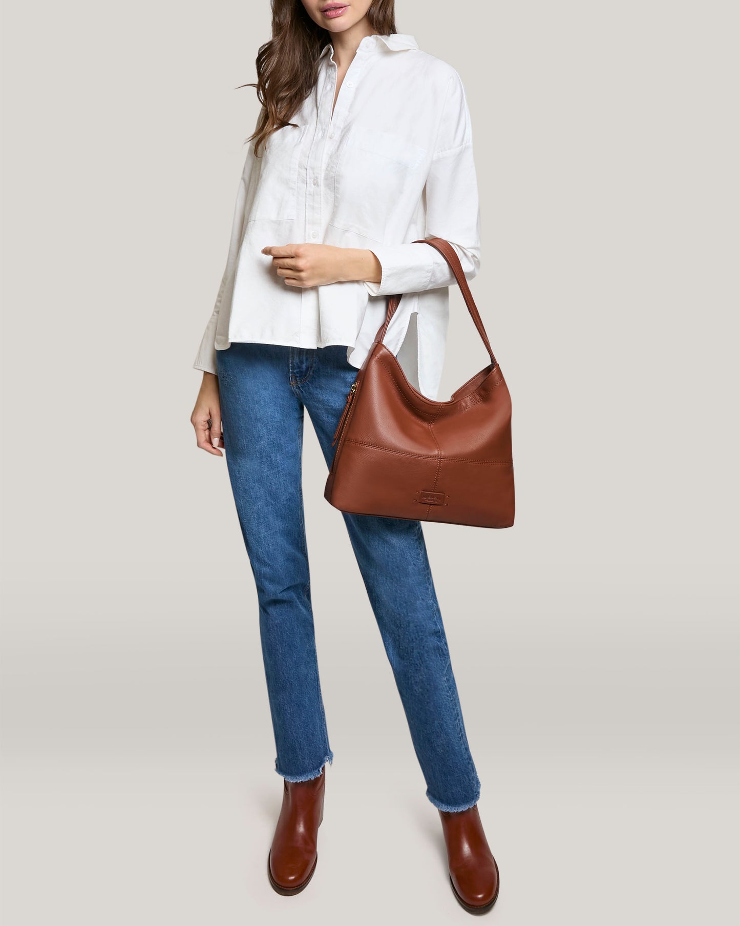 A person in a white shirt, blue jeans, and brown boots holds an American Leather Co. Virginia Hobo with artisanal contrast stitching. The genuine leather's rich tones stand out against the plain, light-colored background.