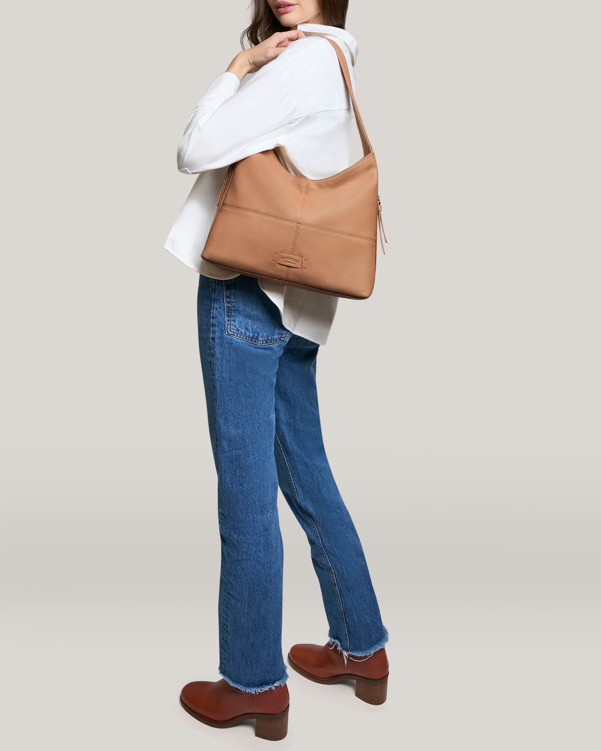 Against a light gray background, a person models the Virginia Hobo by American Leather Co., wearing a white blouse, blue jeans, and brown ankle boots. The satchel features artisanal contrast stitching and is crafted from genuine American leather.