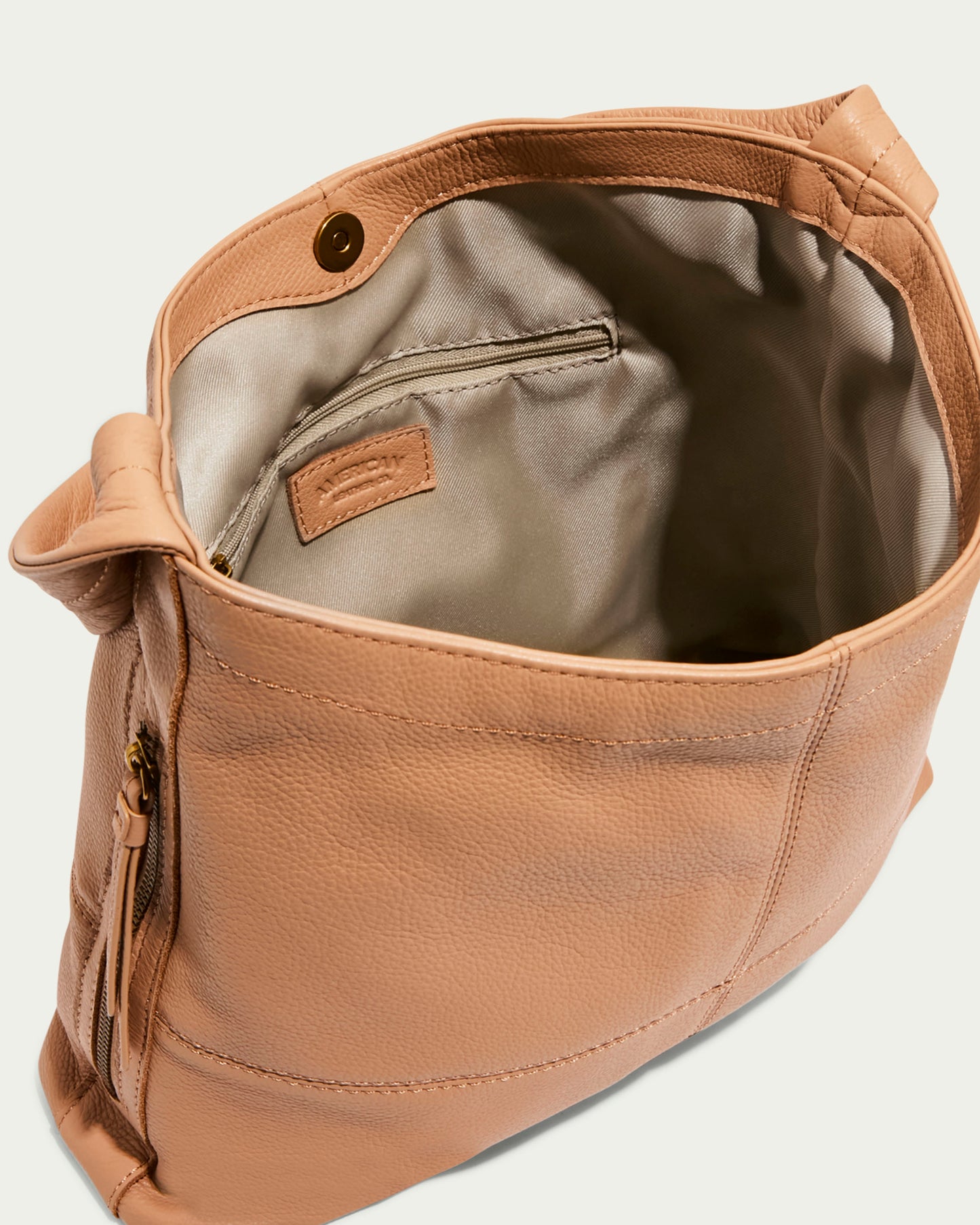 The Virginia Hobo by American Leather Co. is a tan leather satchel with a beige interior, small zippered pocket inside, artisanal contrast stitching, front zippered pocket, and magnetic snap closure. Made from genuine American leather, it combines style and function.