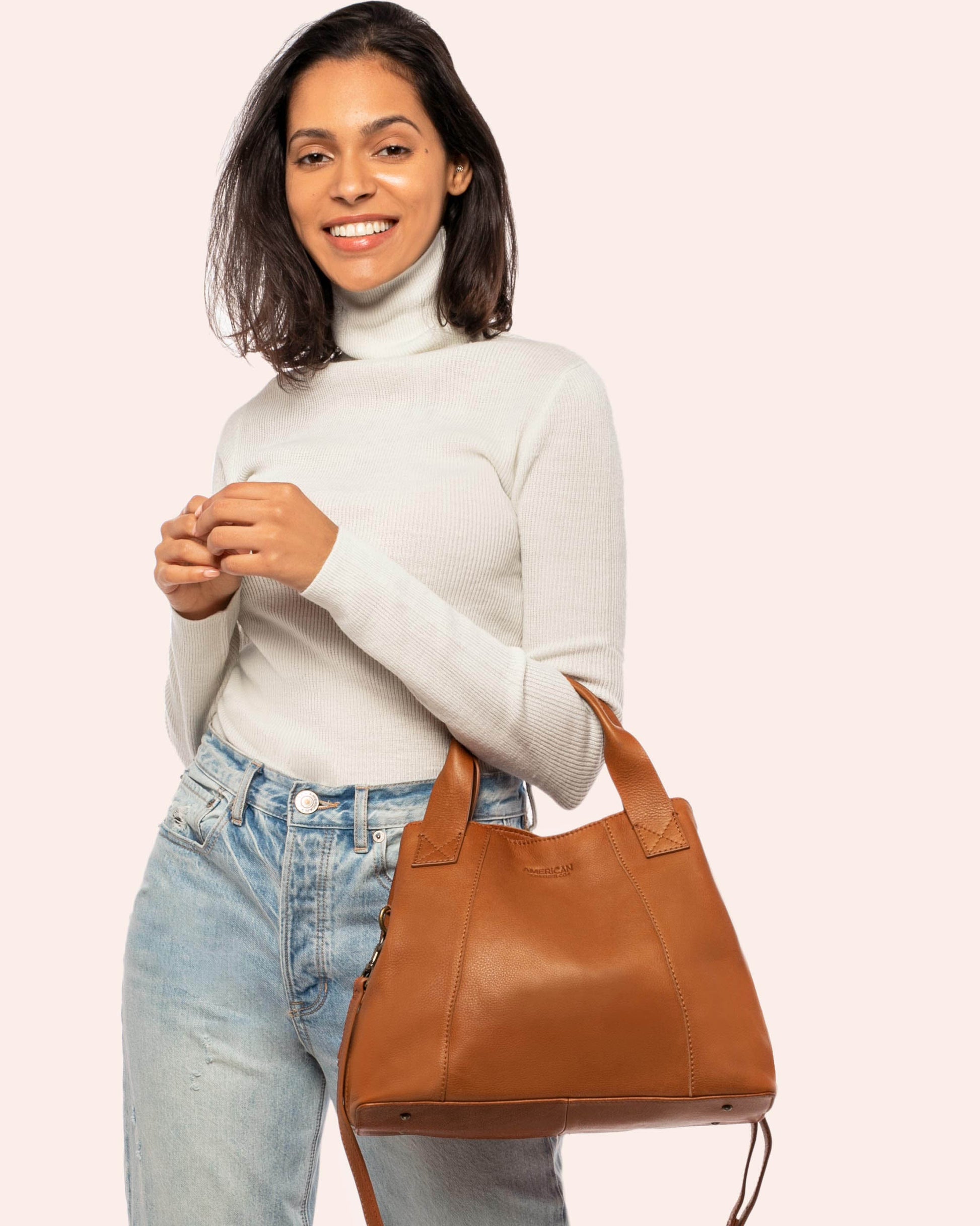 A woman with medium-length dark hair wears a white turtleneck sweater and light blue jeans. She is smiling and holding the Ada Triple Entry Satchel made from Genuine American Leather by American Leather Co. by its handles. The background is a plain off-white color.