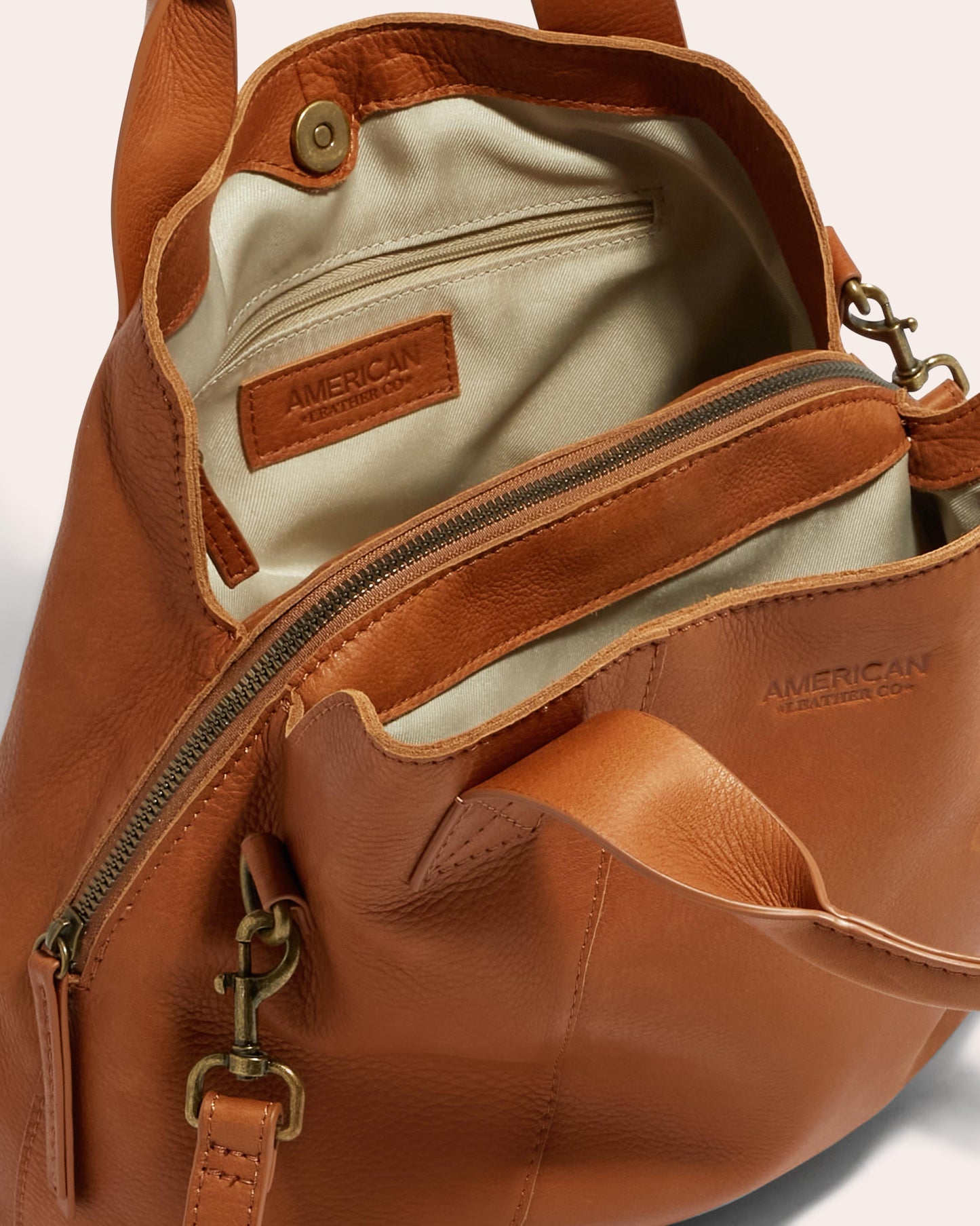 The image showcases the Ada Triple Entry Satchel from American Leather Co., crafted with genuine American leather. The interior is cream-colored, featuring an inner zippered pocket and a stitched leather label. An adjustable crossbody strap, exterior zip pocket, and brass dog clip clasp complete the design.