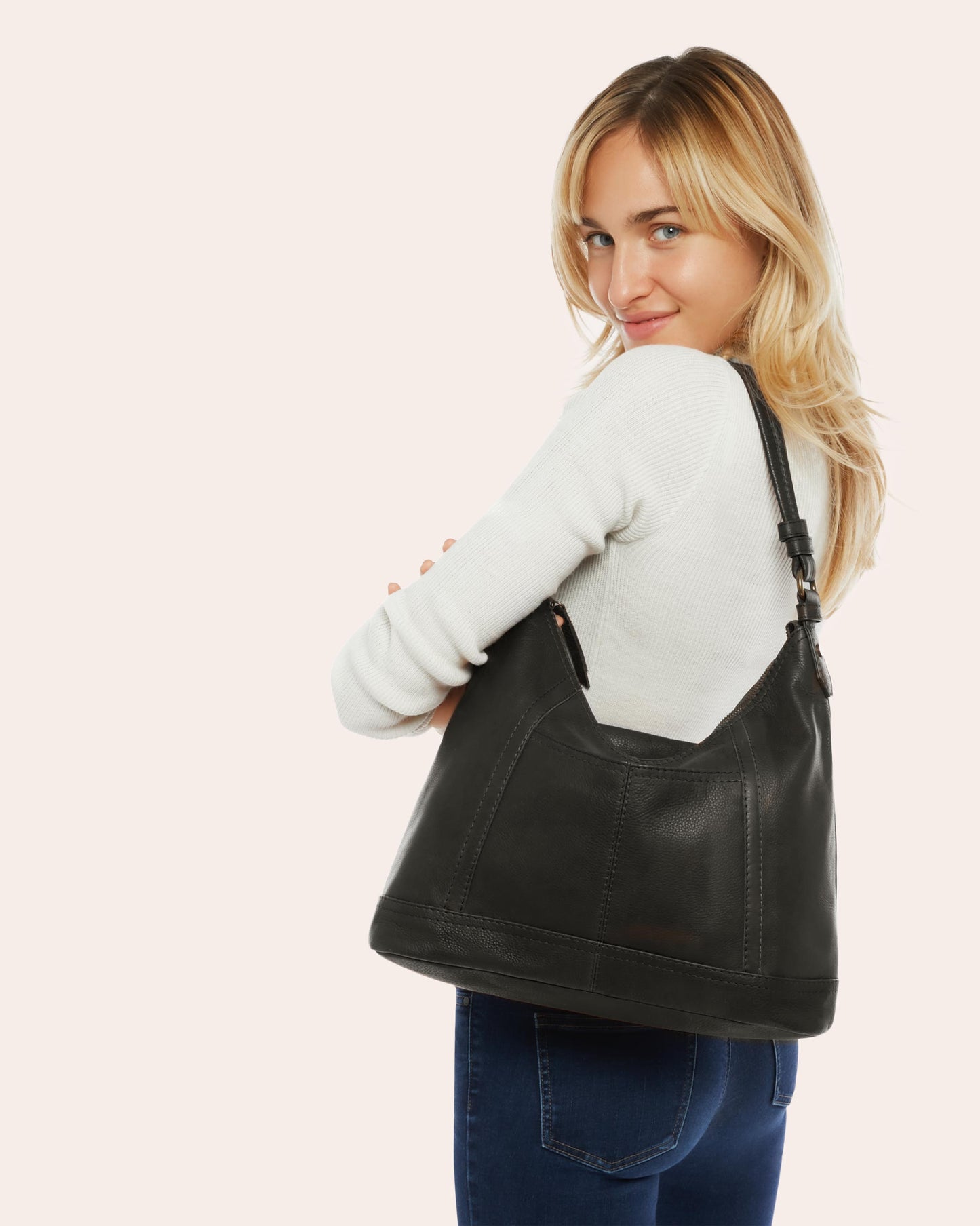 A blonde woman dressed in a white sweater and blue jeans is smiling and looking over her shoulder while carrying the American Leather Co. Hudson Hobo, a black leather shoulder bag featuring antiqued hardware. The background is light pink.