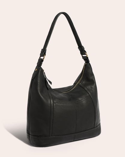 The Hudson Hobo by American Leather Co. is a black leather shoulder bag featuring a single strap and gold-toned, antiqued hardware. This genuine American leather bag has a zipper closure and boasts a slightly slouchy, soft construction. The background is a neutral, pale pink.