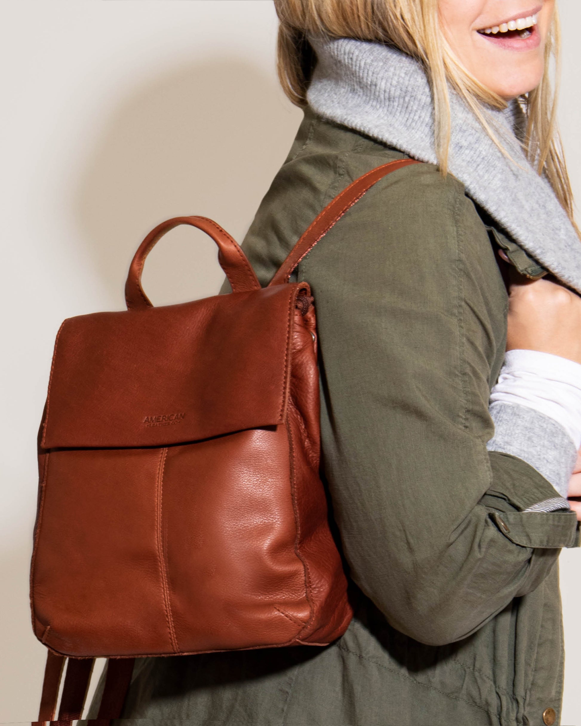 A person wearing a green jacket and a gray scarf is smiling while carrying an elegant Liberty Backpack by American Leather Co. Crafted from fine American leather, this brown backpack features a top handle and adjustable straps for added comfort.