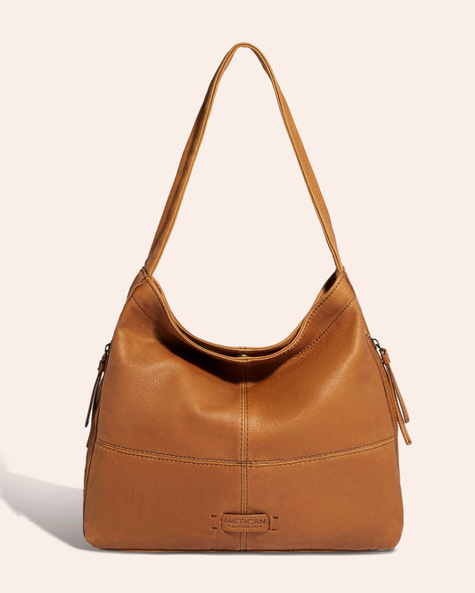 The Virginia Hobo, a medium-sized shoulder bag by American Leather Co., is crafted from Genuine American Leather in brown and features a single, wide shoulder strap. It boasts artisanal contrast stitching and a branded patch at the bottom center, set against a plain, light beige background.